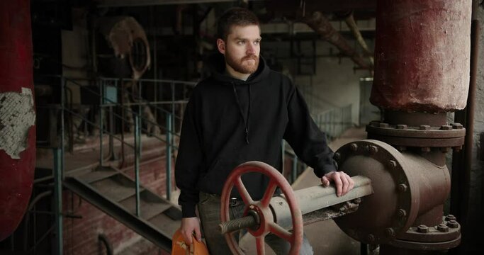 a man with a red beard in a black sweater stands near a shut-off valve in an abandoned factory