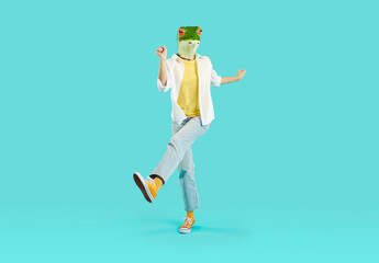 Full length shot of strange woman in funny animal disguise having fun in studio. Full body portrait of happy young girl wearing green frog mask dancing and jumping isolated on turquoise background