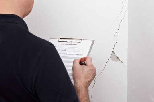 Man with inspection checklist in front of a white wall with a long crack or rip and a piece of plaster missing, rental damage concept.