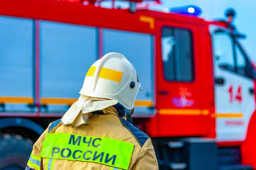 Rear view of a Russian firefighter, rescuer in a protective suit and white helmet fighting a fire....