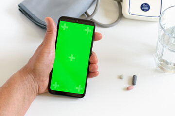 Elder woman holding in wrinkled hands phone with chroma key screen. In the background automatic blood pressure monitor, pills, glass of water. Close-up picture. Selective focus