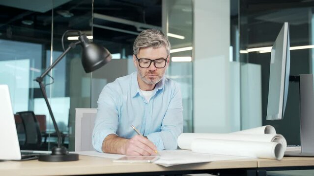 Experienced mature creative designer, architect working in the office at computer desk with drawings. Handsome senior man doing project work sitting at workplace sketching a housing plan building