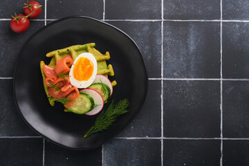 Savory waffles. Breakfast green waffles wild garlic or spinach with egg, tomato, salmon on wooden plate for breakfast on old black tile table background. Light dinner, lunch or breakfast. Top view.