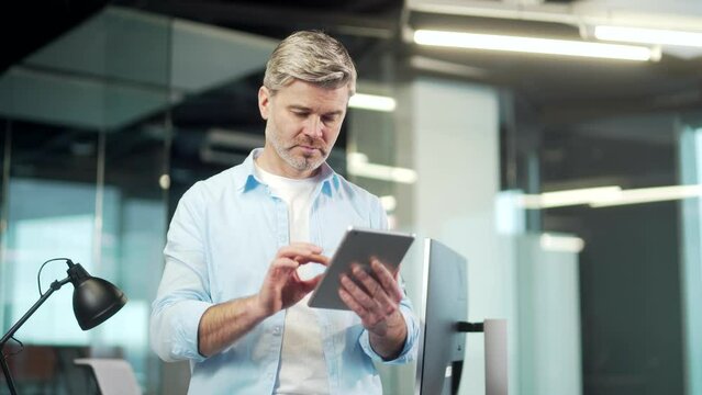 Mature business man uses a digital tablet standing in a modern office indoors. Gray-haired senior male employee worker using Browsing smartphone. Businessman in casual shirt working at workplace