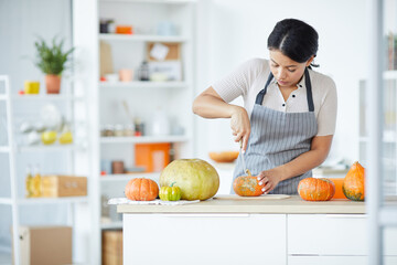 Description: Concentrated young black woman in stripped apron cutting pumpkin on board while carving pumpkin for Halloween in kitchen