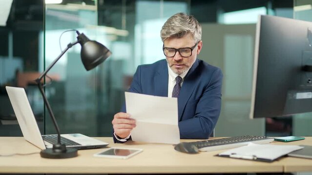 Serious mature businessman in a formal suit examines a document sitting at work in office Gray haired bearded focused business man reads a contract letter or documentation. Lawyer banker on workplace