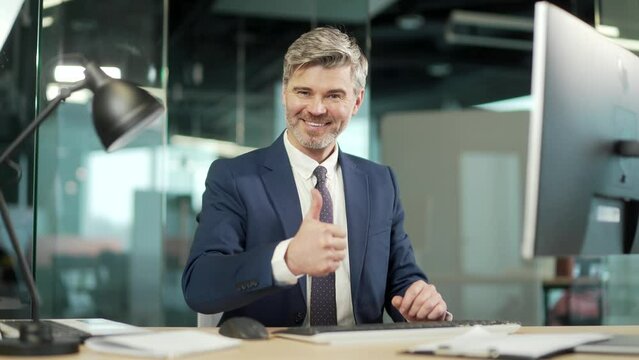 Successful Mature Businessman Wearing Suit Sitting At Desk With Laptop In Office. Portrait of a confident gray hair male in the workplace working at a computer. smiling and looking at camera thumb up