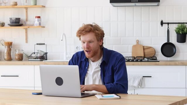 Portrait of excited young Caucasian man with red haired celebrating success while sitting with laptop at home. Happy male freelancer working having good news he receive payment