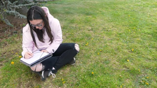 Female writer using a pen to write on a paper sheet while sitting on grass, outdoors video in 4k.