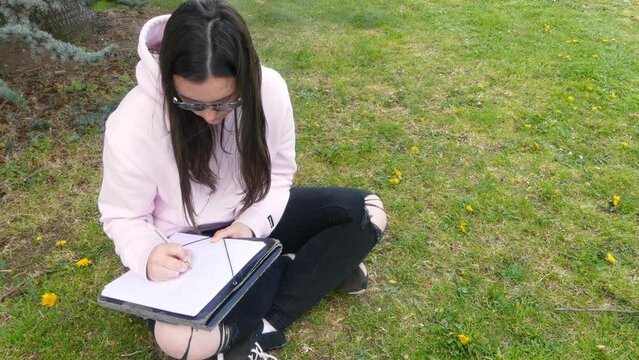Young teenage female writer with glasses writing on a paper sheet using a pen, sitting on grass outside.