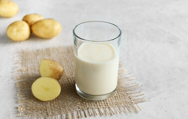 Vegan potato milk in a glass and potatoes on burlap on the wooden background, non-dairy alternative milk,  copy space. Potato milk contains many useful vitamins and minerals vitamin B6, vitamin C.