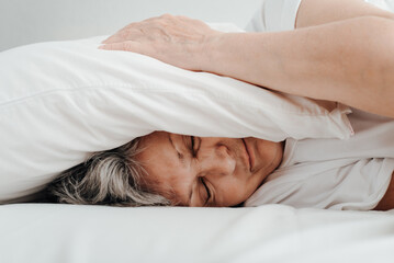Insomnia, headache, depression, female mental health concept. Side view of lonely frustrated senior woman with closed eyes covering her head with pillow while lying in bed in bedroom, close-up
