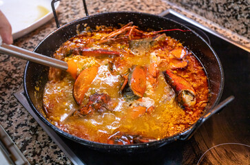 Induction cooking of a lobster rice paella. Metal and plastic ladle stirs the contents of the paella prepared at home with care and love.