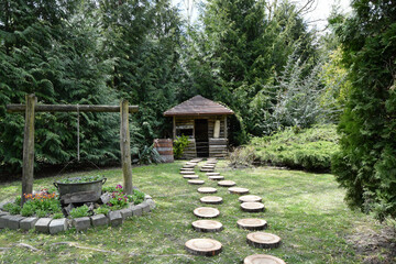 Beautiful view of a small wooden house surrounded by green trees. Path of round pieces that wood taking the place of the alley. Garden decorations