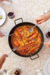 Four hands with their cutlery want to start a rice with lobster served in a black paella on a prepared table.