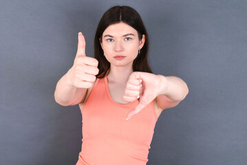 Caucasian woman wearing orange T-shirt over grey wall showing thumbs up and thumbs down, difficult...