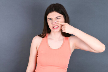 Caucasian woman wearing orange T-shirt over grey wall pointing unhappy to pimple on forehead, ugly...