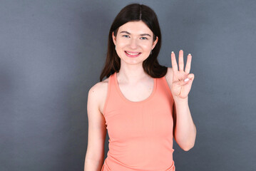 Caucasian woman wearing orange T-shirt over grey wall showing and pointing up with fingers number three while smiling confident and happy.