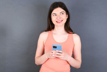 young beautiful Caucasian woman wearing orange T-shirt over grey wall hold telephone hands read good youth news look empty space advert