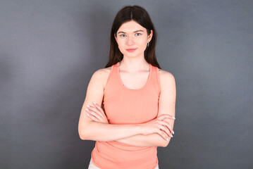Obraz na płótnie Canvas Self confident serious calm young beautiful Caucasian woman wearing orange T-shirt over grey wall stands with arms folded. Shows professional vibe stands in assertive pose.