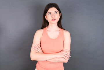 Fototapeta na wymiar Charming thoughtful young beautiful Caucasian woman wearing orange T-shirt over grey wall stands with arms folded concentrated somewhere with pensive expression thinks what to do