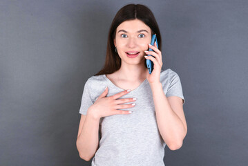 Smiling  young beautiful Caucasian woman wearing white T-shirt over grey wall  talks via cellphone, enjoys pleasant great conversation. People, technology, communication concept
