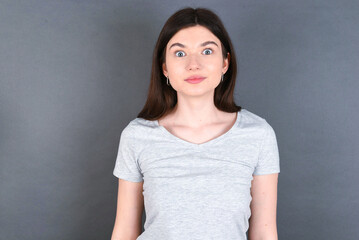 Obraz premium Stunned young beautiful Caucasian woman wearing white T-shirt over grey wall stares reacts on shocking news. Astonished girl holds breath