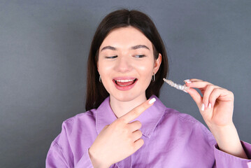 Young smiling showing perfect teeth caucasian woman wearing pink shirt standing over gray background, holding an invisible aligner. Dental healthcare concept. 