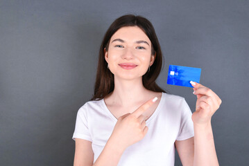 Beautiful caucasian girl standing over gray background and wearing white T-shirt, holding a credit card and pointing at it. Finance and online shopping concept.