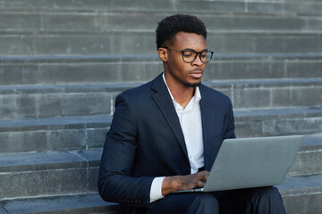 Serious concentrated young African-American man in suit sitting on stairs and typing on laptop while working on report out of office - obrazy, fototapety, plakaty