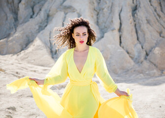 beautiful brunette woman with curly hair walking in long yellow dress on the beach in mountains