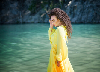 beautiful brunette woman with curly hair walking in long yellow dress on the beach in mountains