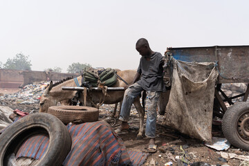 Hopeless African young boy sitting alone on a handcart with a donkey, collecting waste and garbage...