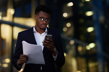 Serious busy young black man in eyeglasses standing against office building at night and holding papers while checking information on internet