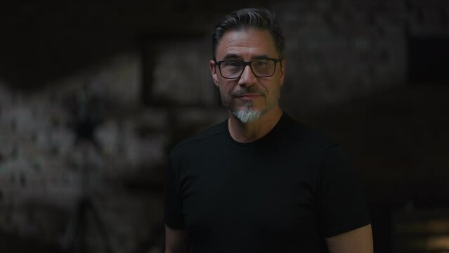 Portrait of confident middle aged man in a dark industrial room wearing glasses. Low light, copy space .