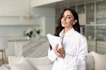 Brunette woman in white clothes holding papers standing indoors in living room in apartment, in design interior. Freelancer, student or business woman at home office, education or remote work.