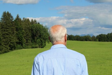 Älterer Mann mit grauen Haaren im blauen Hemd schaut in die hügelige Landschaft im Allgäu, Bayern