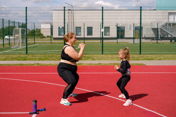 Young mother and skinny daughter play sports together, doing squat exercises on sports field. Plump...