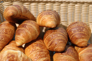 Big pile of multiple big and crunchy breakfast croissants in a wooden basket. Photographed outdoors during a sunny summer day. Delicious Parisian style breakfast bread. Closeup color image.