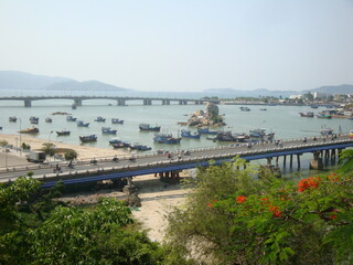 View of a road bridge over a river in a tropical country. Cars and motorbikes are driving along the bridge. Boats are in the river.