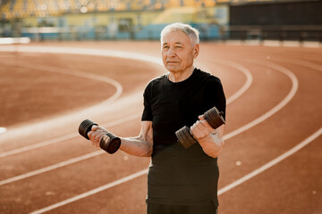 Close up portrait of an elderly man in black sportswear. Senior man exercising with dumbbells at the stadium. Sport and lifestyle content.