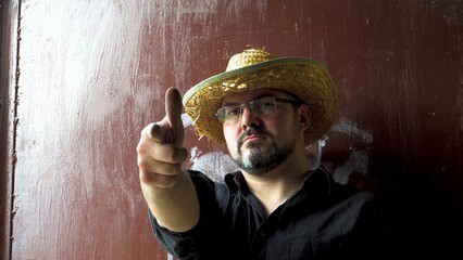 An adult man in a straw hat shoots from his finger. He folds his hands with a pistol. Bearded and wearing glasses. Close-up. Military man. Farmer. Cowboy. On a dark background.