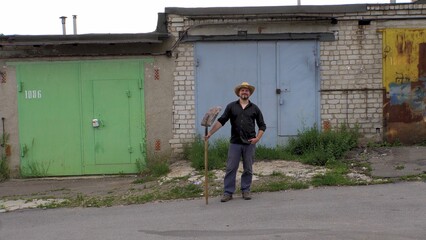 A man in a straw hat holds a shovel against the backdrop of garages. Dressed in a black shirt and pants. Bearded. warm day. worker. builder. Illegal migrant. Searching for a job