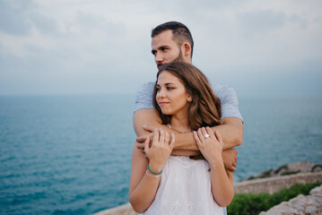A Hispanic man is hugging from behind his brunette girlfriend in the park in Spain in the evening. A couple of tourists are staring at the sea on a date at the sunset in Valencia.