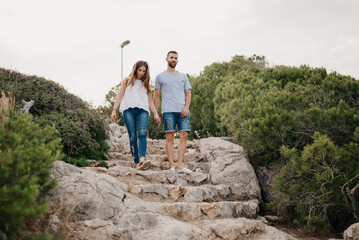 A Hispanic man is helping his wife descend the ancient stone stairs in the highland park in Spain. A couple of tourists are enjoying hiking with each other on a date at sunset in Valencia.