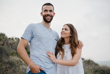 A Hispanic man is hugging his happy girlfriend in the highland park in Spain in the evening. A couple of tourists are enjoying each other near the sea on a date at sunset in Valencia.