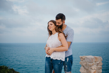 A Hispanic man is hugging from behind his brunette wife in the park in Spain in the evening. A couple of tourists are enjoying each other near the sea on a date at the sunset in Valencia.