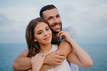 A close photo of a Hispanic man who is hugging from behind his brunette wife in the park in Spain in the evening. A couple of tourists are enjoying the sea view on a date at the sunset in Valencia.