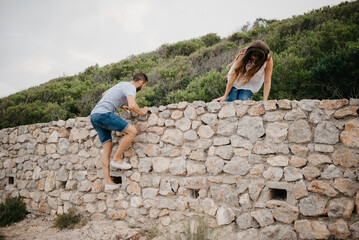 A Hispanic man and his girlfriend are climbing over the ancient stone wall in a highland park in Spain. A couple of tourists are enjoying hiking with each other on a date at sunset in Valencia.