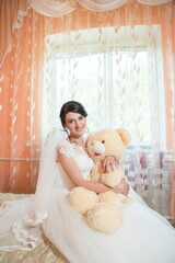 Fototapeta na wymiar The bride at a wedding with a toy bear sits on a bed by the window.